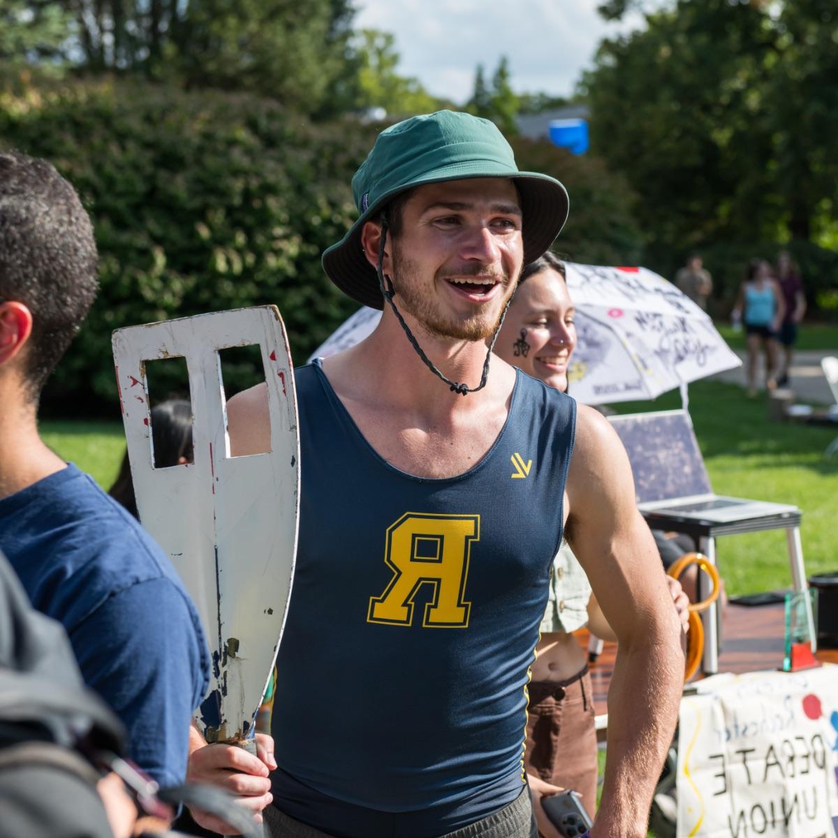 University of Rochester athlete wearing a bucket hat and University branded tank top holds an oar